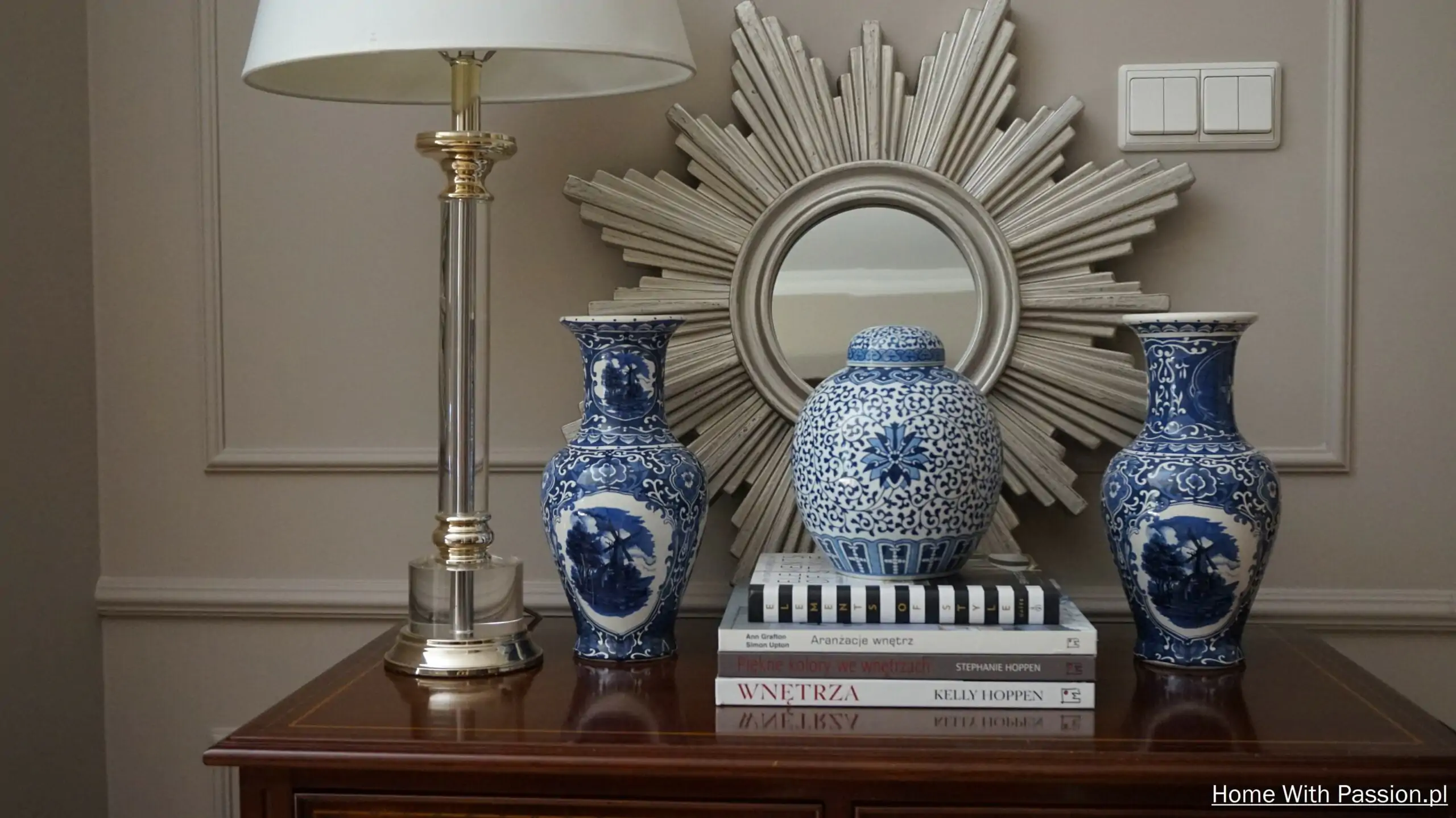 Biało-niebieskie wazony i ceramiczny pojemnik na książkach pod dekoracyjnym lustrem – elegancka aranżacja komody w stylu hampton